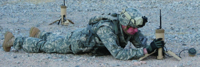 A soldier in full combat gear lies prone on a gravelly surface while deploying a tactical ground sensor. Another sensor stands deployed in the background.